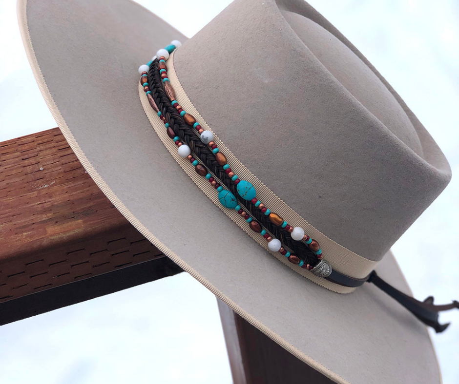 This trendy hatband features a seven strand braid made of genuine horse hair, adorned with coppery metal beads, white and turquoise howlite, and freshwater pearls. It is completed with our signature sunburst conchos, leather tassels, and a horse hair knot for secure fit. Designed to fit most hats, it is available at our Smyrna, Tn shop. Every piece is carefully handcrafted in Montana using horse hair from various sources including Argentina, Mongolia, and Canada.